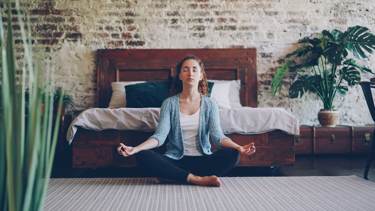 Beautiful young lady is sitting in lotus position on bedroom floor and enjoying meditation, harmony and peace. Modern loft style houses, healthy people and rest concept.